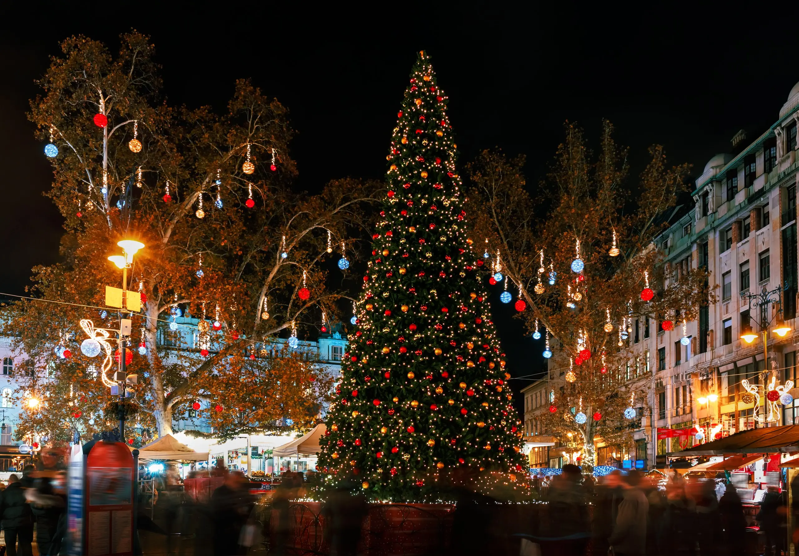 A Budapest Christmas market at dusk, a tall decorated tree lit above the stalls and the crowd.