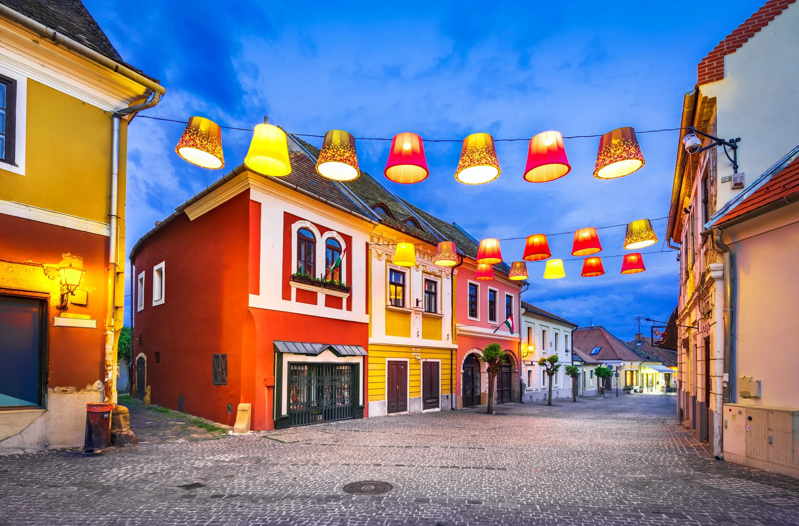 A cobbled street in Szentendre at dusk, ochre and red houses under a string of yellow lampshades.